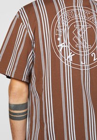 Hnědá a bílá vertikálně pruhovaná košile s bílým kruhovým logem a částečným tetováním černých pásků na předloktí.