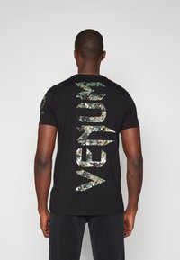 T-shirt noir à manches courtes avec un grand logo en motif camouflage au dos. Tissu doux avec un col rond et une coupe décontractée.