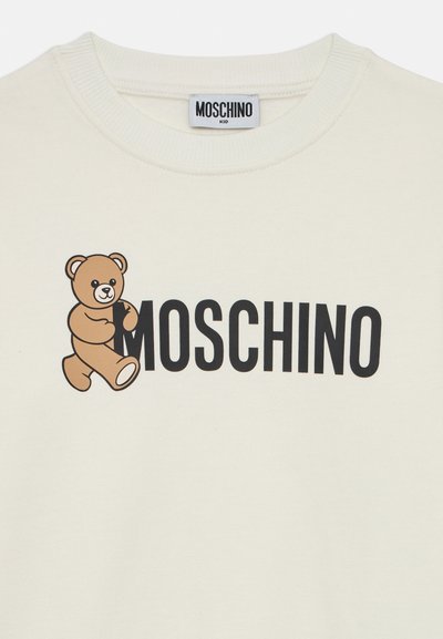 Hvit bomullsgenser med et brunt bjørnemotiv til venstre og "MOSCHINO" tekst i fete svarte bokstaver under. Rund halsdesign.
