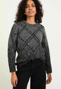Frau trägt einen grauen und schwarzen, flauschigen Strickpullover mit Rautenmuster, kombiniert mit hochgeschnittenen schwarzen Hosen, stehend vor einem schlichten Hintergrund.