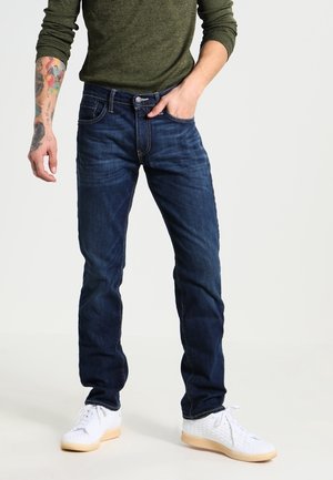 Homme portant un jean bleu foncé, des baskets blanches et une chemise à manches longues vert olive, avec l'avant-bras gauche tatoué visible.