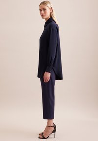 Camicia blu navy a maniche lunghe con dettagli in bottone, abbinata a pantaloni blu navy su misura e sandali neri con tacco, mostrata in vista profilo.