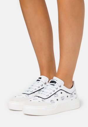 Jalat yang memakai sepatu sneaker MCM rendah putih dengan logo hitam dan sol tebal putih di latar belakang terang yang polos.