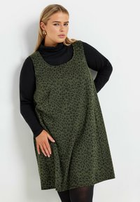 Vihreä oliivinvihreä hihaton mekko mustalla leopardikuosisella, valmistettu pehmeästä kankaasta. Pukeutuu mustan poolopaidan päälle, jossa on pyöreä pääntie ja rento istuvuus.