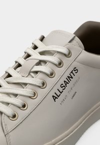 Κοντινό πλάνο μπεζ αθλητικού παπουτσιού AllSaints με λευκά κορδόνια και ιαπωνικό κείμενο πάνω από τη λέξη "Λονδίνο" στην πλευρά του παπουτσιού.