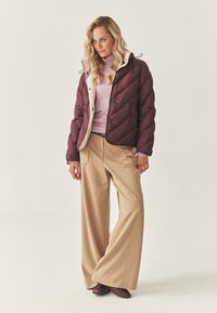 Bordeaux pufferjakke med en cremefarvet inderside, parret med en lys pink turtleneck og beige vidde-bukser. Brune sko fuldender outfittet.