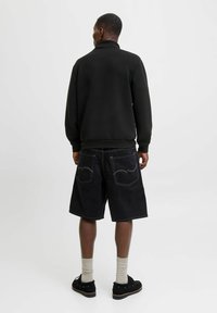Mand står med ryggen til iført en sort sweatshirt, lange sorte shorts med synlig hvid syning, beige sokker og sorte sko.