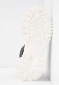 Semelle de botte noire avec une épaisse semelle blanche, positionnée verticalement contre un mur et un sol blancs.