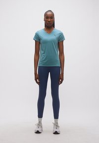 Γυναίκα που στέκεται όρθια φοράει μπλουζάκι Puma σε βεραμάν χρώμα με V λαιμόκοψη, μπλε σκούρο κολάν, άσπρες κάλτσες και άσπρα αθλητικά παπούτσια μπροστά σε απλό φόντο.