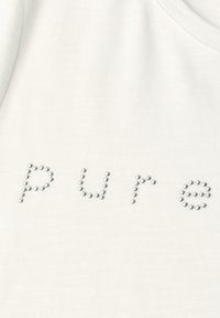 Gros plan sur un tissu blanc avec le mot « Pure » épelé en petites clous ronds argentés disposés en minuscules.