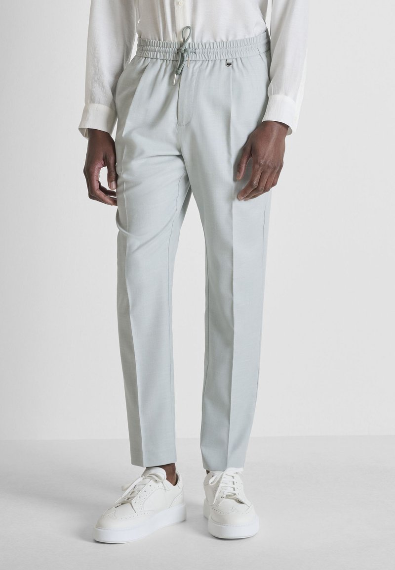 Hombre con pantalones gris claro con cordón, camisa blanca y zapatillas plataforma blancas, de pie sobre un fondo liso.