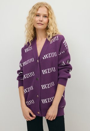 Vrouw met golvend blond haar, die een losse paarse vest met knopen en witte "BIZUU" tekst draagt, staande tegen een effen lichte achtergrond.