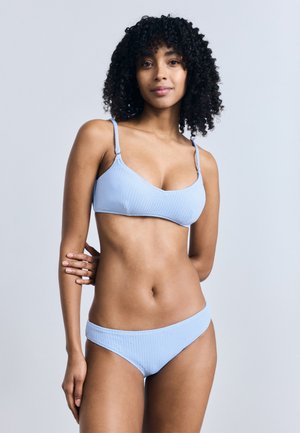 Kvinde med krøllet hår iført en lyseblå tekstureret bikini, stående med den ene hånd på hoften mod en ensfarvet lys baggrund.