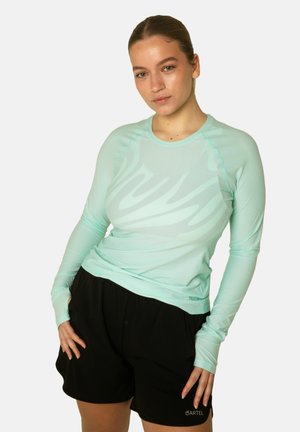 Kobieta w miętowo-zielonym długim rękawie i czarnych szortach z logo "CARTEL", pozująca z rękami opartymi na biodrach.