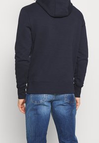 Osoba v tmavě námořnické mikině s kapucí a modrých džínách, zobrazená zezadu na jednobarevném světlém pozadí.