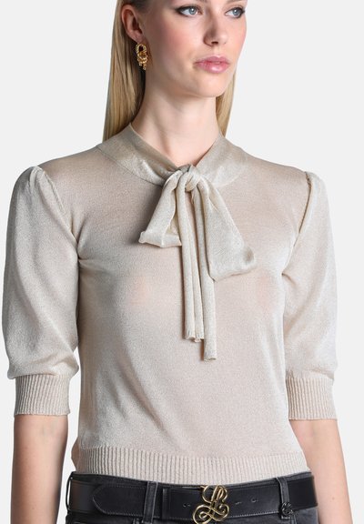 Femme portant un chemisier beige clair à manches courtes avec un col à nœud papillon et une ceinture noire avec une boucle décorative dorée.