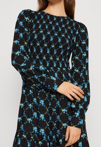 Musta kukkamekko, jossa on sinisiä ruusuja ja vihreitä yksityiskohtia, pitkät puffihihat, teksturoitu kangas ja kerroksellinen hame. Napitus yksityiskohtana hihassa.