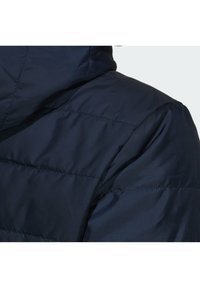 Pohled zblízka na rameno a kapuci tmavě modré prošívané bundy s viditelnými vodorovnými stehy.