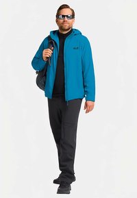 Blauwe jas met ritssluiting over een zwart shirt, gecombineerd met een zwarte broek en schoenen. Accessoire: zwarte tas. Buitenkleding afgebeeld tegen een neutrale achtergrond.