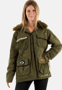 Parka verde oliva con cuello de piel sintética, bolsillos adornados y parches. Presenta tachuelas metálicas, detalle de ojo bordado y motivos de estrellas.