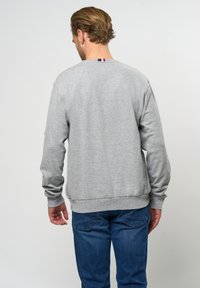 Grå hettegenser med lange ermer, ribbestrikkede mansjetter og søm. Baksiden har en liten, horisontal tri-farge aksent nær halsen. Brukt med blå jeans.