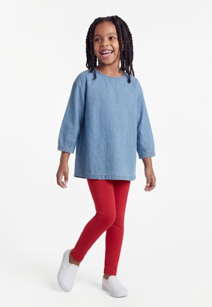 Lachend meisje met gevlochten haar, gekleed in een blauw shirt met lange mouwen, rode legging en witte instapschoenen, lopend tegen een witte achtergrond.