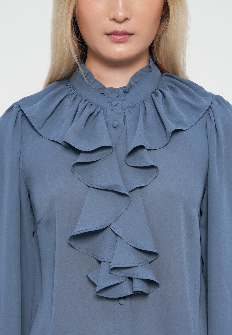 Blaue Bluse mit hohem, rüschendem Kragen und herabfallenden Vorderüsche; aus glattem Stoff mit Knopfdetails am Hals.