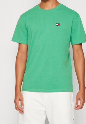 Mann trägt ein grünes T-Shirt mit kurzen Ärmeln und kleinem Tommy Jeans-Logo auf der Brust sowie weiße Shorts.