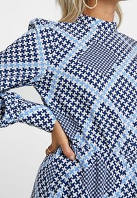 Blå och ljusblå mönstrad klänning med geometriska mönster, hög halsringning och långa ärmar. Tyget har en jämn textur.
