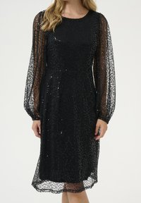 Robe noire avec manches longues transparentes, corsage cintré et motif texturé avec des sequins sur toute la surface. Ourlet à hauteur des genoux.