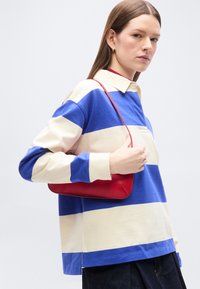 Femme portant un chemisier rayé bleu et blanc à manches longues, tenant un sac à bandoulière rouge, de profil avec une expression neutre.