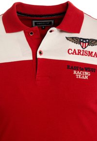 Rød polo skjorte med hvid panel, der har broderi med teksten "CARISMA," et ørnedesign og "EAST to WEST RACING TEAM." Akrylmateriale.