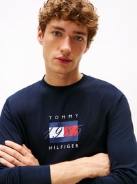 Mornarska majica z dolgimi rokavi z osrednjo grafiko, ki vsebuje "TOMMY" in "1985", v rdeči, beli in modri barvi. Mehka bombažna tkanina.