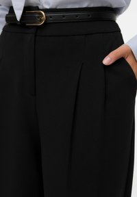 Detailní záběr na černé plisované kalhoty s černým páskem a rukou zasunutou v pravé kapse, doplněné o světlou košili.