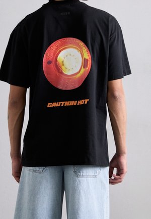Personne portant un T-shirt noir avec un grand graphisme rouge et jaune « Attention Chaud » dans le dos, associé à un short en jean bleu clair.