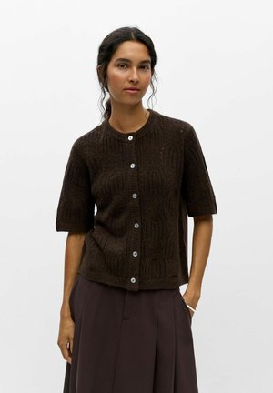 Femme aux cheveux foncés portant un cardigan en maille brun foncé à manches courtes avec des boutons et un pantalon brun foncé ample, debout devant un fond uni.