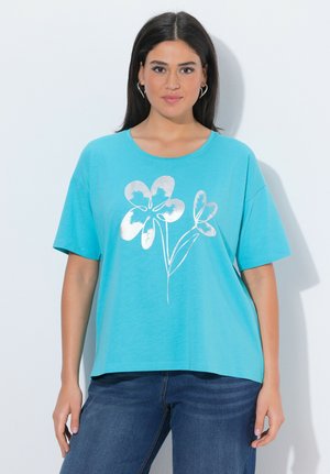 Kvinde med langt mørkt hår iført en turkis t-shirt med et sølvfarvet blomsterdesign og blå jeans, stående foran en hvid baggrund.