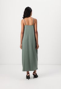 Green, sleeveless maxi dress with thin straps, made of soft fabric, featuring a straight cut and flowing silhouette. Worn with black heeled sandals.