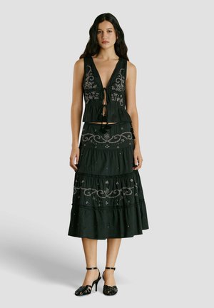 Naine, kes kannab musta varrukateta toppi ja sellele sobivat hõbedaste broderiidega midi-seelikut, mustade kontsakingadega, seisab tavalise tausta ees.