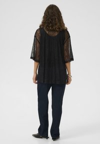 Femme aux cheveux bouclés portant un top en dentelle noire transparente, un jean droit bleu foncé et des escarpins à talons kitten noirs, se tenant de dos.