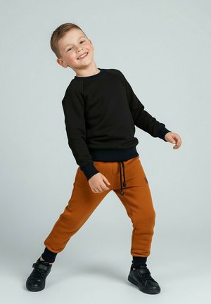 Uśmiechnięty młody chłopiec stoi z rozstawionymi nogami, ubrany w czarną bluzę, rdzawobrązowe spodnie dresowe i czarne buty sportowe na jednolitym, jasnoszarym tle.