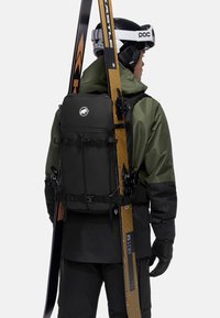 Svart ryggsekk med justerbare stropper, flere spenner, en logo foran, og to ski sikret ved siden av. Brukt med en grønn jakke.