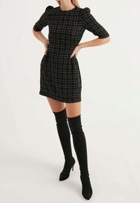 Schwarzes, tailliertes Kleid mit Karomuster, kurzen Puffärmeln, rundem Ausschnitt und zwei seitlichen Taschen. Kombiniert mit schwarzen, über dem Oberschenkel hohen Stiletto-Stiefeln.