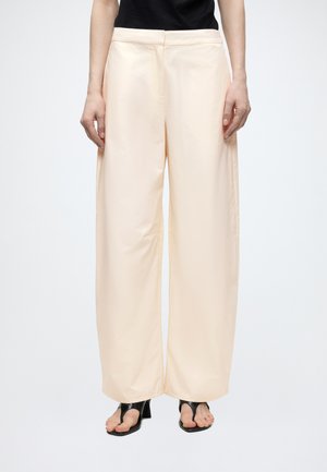 Pantalon crème à jambes larges porté par une personne en sandales à talons ouvertes noires et un haut noir, debout devant un fond blanc uni.