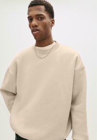 Hanorac oversize bej, confecționat dintr-un material moale, cu guler rotund și umeri lăsați, având o textură ușor ștampilată și un design minimalist.