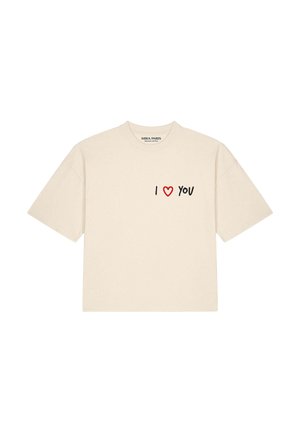 Frau mit beigen Sonnenbrillen und einem cremefarbenen oversized T-Shirt mit dem Text "I heart you" und beigen Hosen vor weißem Hintergrund.