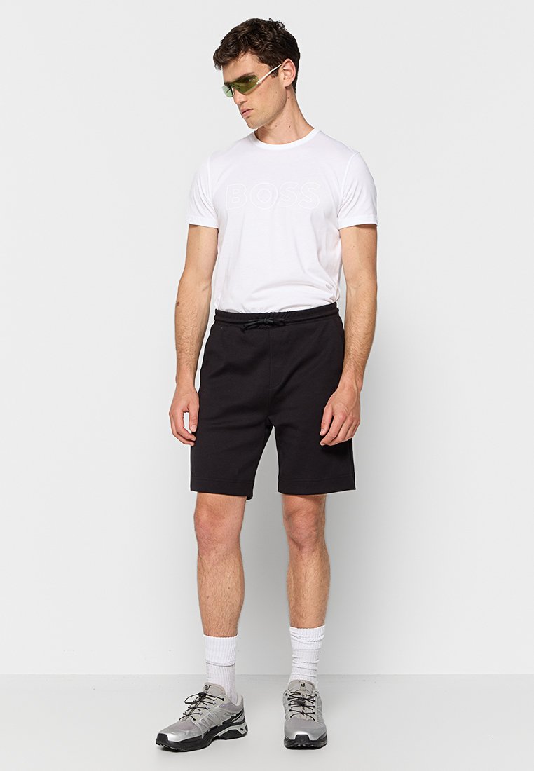 Czarne sportowe szorty z elastycznym paskiem i sznurkiem, zestawione z białą koszulką z wytłoczonym logo „BOSS” oraz szarymi butami sportowymi.