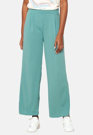 Pantalons évasés aqua en tissu doux avec une texture lisse et un design plissé, associés à des baskets blanches.