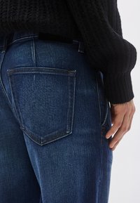 Tumesinised teksapüksid tekstuurse viimistlusega, millel on kaks tagataskut ja kontrastõmblused. Vöökohal on bränditud silt.