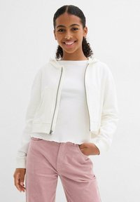 Felpa bianca corta con zip e cappuccio, abbinata a un top bianco a coste e pantaloni in velluto a coste rosa chiaro, con orlo grezzo.